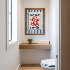 May include: A framed wall art print with the text "Rootin' & Tootin'" and a cowboy illustration. The artwork is in a wooden frame and hangs above a wooden shelf with a small plant and towels. The background is a white wall.