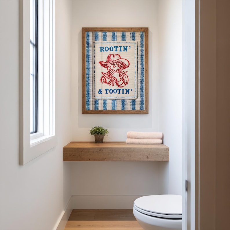 May include: A framed wall art print with the text "Rootin' & Tootin'" and a cowboy illustration. The artwork is in a wooden frame and hangs above a wooden shelf with a small plant and towels. The background is a white wall.