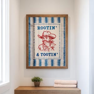 Może przedstawiać: Oprawiona grafika z ilustracją kowboja i napisem "ROOTIN' & TOOTIN'". Grafika jest w drewnianej ramie, z niebiesko-białym pasiastym tłem. Kowboj ma na sobie kapelusz i zakrywa usta.