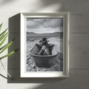 K&ouml;nnte beinhalten: Ein Schwarzwei&szlig;foto einer Person, die einen Cowboyhut und Stiefel tr&auml;gt und in einer Badewanne in der W&uuml;ste sitzt.