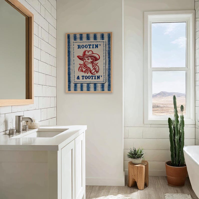 Framed “Rootin’ & Tootin’” red and blue cowboy wall art hanging above a sink in a bright, neutral-toned bathroom with soft natural light and cozy, rustic decor.
