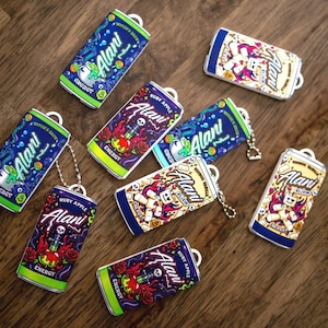 May include: A collection of rectangular keychains shaped like Alani energy drink cans. The designs include Witch's Brew with green accents, Ruby Apple with red roses and skulls, and Voodoo Vanilla featuring a doll graphic. Each has a metal chain.