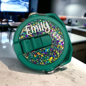 Peut inclure: Un couvercle de bouteille d'eau vert d'eau avec le nom "Emily" en lettres blanches. Le couvercle comporte une section circulaire transparente remplie de paillettes colorées. Le couvercle a une fermeture à rabat et une charnière argentée.