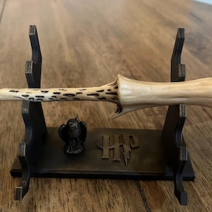 May include: A light-colored, textured wand rests horizontally on a black display stand. The stand features a small owl figurine and the letters HP in a stylized font. The wand has a tapered shape with dark, pitted markings along its thinner end.