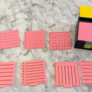 May include: A set of pink sticky note pads with different grid patterns. The pads are in a black holder with a yellow sticky note on top.