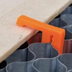 May include: A bright orange L-shaped plastic clip securing a flat wooden board to a metallic honeycomb-patterned laser cutter bed. The clip features a flat top edge and a vertical stem that fits into the grid structure of the metal base.