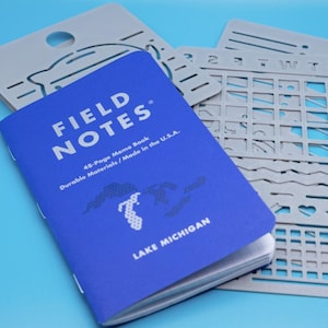 May include: A blue Field Notes 48-page memo book with white text reading Field Notes, Durable Materials / Made in the U.S.A., and Lake Michigan, featuring a graphic of the lake, accompanied by two grey plastic stencils with various cutouts.