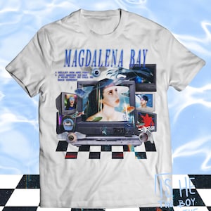 Puede incluir: Camiseta blanca con el nombre de la banda "MAGDALENA BAY" en azul. El diseño muestra a una mujer dentro de un televisor retro, con otras pantallas y el texto "IS HE THE BOY FROM THE VIDEO".