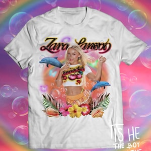 Puede incluir: Camiseta blanca con un gráfico de Zara Larsson y el texto "Zara Larsson". El diseño incluye delfines, flores y el texto "connected". La camiseta tiene un estilo pop art colorido, con un arcoíris y burbujas en el fondo.