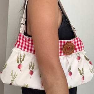 Puede incluir: Bolso de hombro con fondo blanco y estampado de rábanos rojos con hojas verdes. La parte superior del bolso es de cuadros rojos y blancos, con un gran cierre de botón de madera. El bolso tiene una correa blanca.