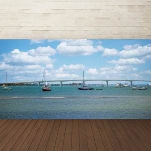 May include: A panoramic view of a bridge over a body of water with sailboats and motorboats. The sky is blue with white clouds.