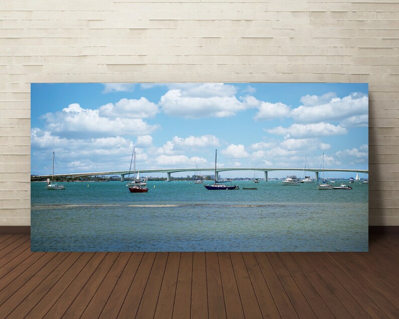 May include: A panoramic view of a bridge over a body of water with sailboats and motorboats. The sky is blue with white clouds.