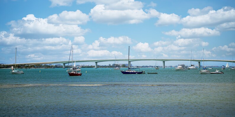 May include: A view of a bridge over a body of water with several sailboats docked. The sky is blue with white clouds.
