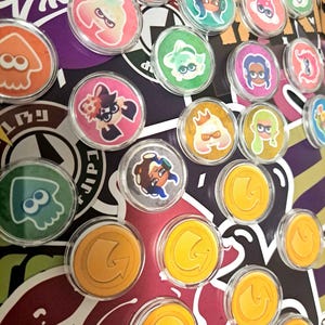 May include: A collection of clear, circular buttons featuring colorful cartoon characters and gold-colored coins with arrows. The buttons have a glossy finish and are arranged on a patterned background with purple, red, and white elements.