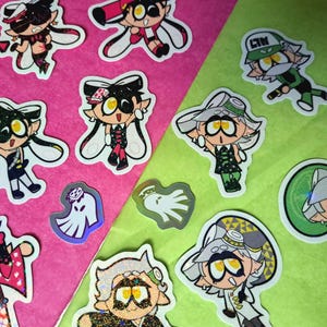 May include: A collection of die-cut stickers featuring cartoon characters with large yellow eyes and various outfits, including some with glittery finishes and two small ghost-shaped holographic stickers, arranged on a pink and green background.