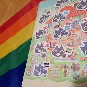 Puede incluir: Un cuaderno con una colorida ilustración de una escena de pueblo y pegatinas de un gato con banderas del orgullo. Una bandera arcoíris es visible en el fondo. Las pegatinas tienen un efecto holográfico.