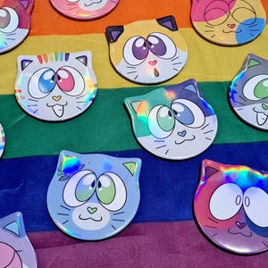 May include: A selection of cat-shaped holographic stickers in various colours. Each sticker has a unique cat face design with large eyes and a small heart-shaped nose. The stickers are displayed on a rainbow-coloured background.