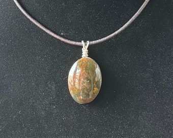 May include: A brown leather cord necklace with a silver pendant featuring a green, brown, and white oval-shaped stone.
