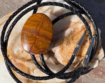 Può includere: Una cravatta a bolo con una pietra ovale lucidata che mostra striature marroni e arancioni. La pietra è montata su un cordino in pelle intrecciata nera con punte metalliche argentate.