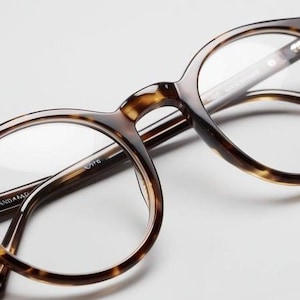 Könnte beinhalten: Schildpatt-Brille mit runden Gläsern und klaren Gläsern. Die Brille hat eine dezente braune und goldene Farbe.