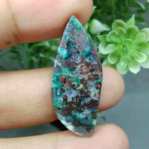 Peut inclure: Une pierre précieuse en forme de marquise présentant un motif tacheté de bleu sarcelle profond, de vert vif et d'inclusions brun cuivre métallique. La pierre a une surface lisse et polie avec une qualité translucide et des textures minérales variées.