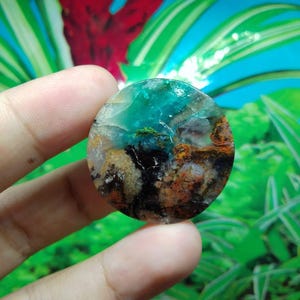 May include: A circular gemstone, polished to a high sheen, displaying a range of colours. The upper section is a vivid teal, blending into brown, black, and orange tones. The stone is held in a hand, with a blurred green and blue backdrop.
