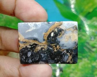 Puede incluir: Una gema rectangular con un patrón único de colores negro, gris y marrón. La piedra se sostiene entre los dedos, mostrando su superficie pulida y variaciones naturales. El diseño de la gema se asemeja a un paisaje.