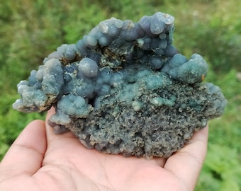 May include: A green and gray agate geode with a rough, textured surface. The geode is filled with small, round, blue-green crystals.