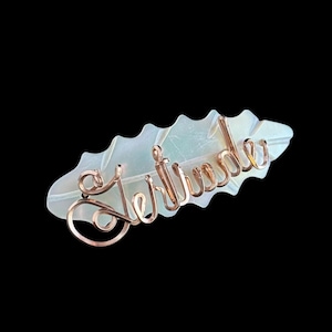 Peut inclure: Une barrette à cheveux avec le nom "Gertrude" en fil de cuivre. Le nom est fixé sur une base nacrée en forme de feuille. Le fond est noir.