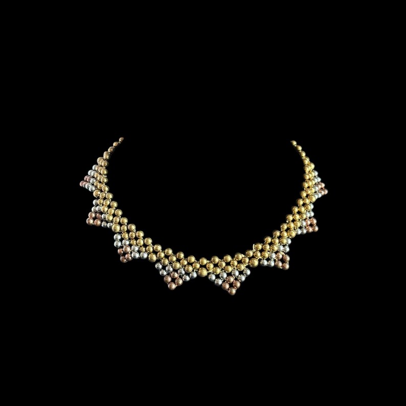 May include: A necklace with a scalloped design, made from gold, silver, and rose gold beads. The beads are arranged in a repeating pattern. The necklace is set against a black background. The necklace's design is eye-catching and textured.
