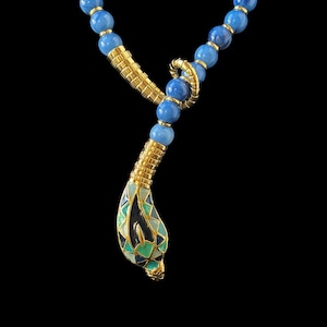 Puede incluir: Un collar con cuentas azules redondas intercaladas con espaciadores cilíndricos dorados. El colgante es un diseño de cabeza de serpiente dorada decorado con patrones geométricos de esmalte azul, verde y blanco, unido al collar.