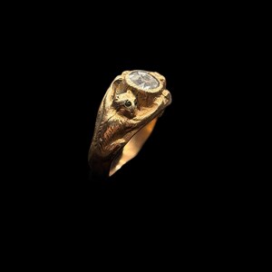 May include: A gold ring featuring a squirrel design. The ring has a clear, round gemstone set in a decorative bezel. The squirrel is sculpted on the side of the ring, adding a unique, nature-inspired element to the jewelry.