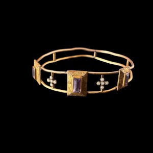 Peut inclure: Un bracelet jonc en métal doré orné de montures rectangulaires avec des pierres rectangulaires violettes et de petits accents en forme de croix agrémentés de minuscules perles blanches. Les surfaces métalliques présentent des gravures.