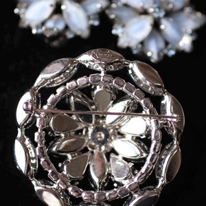 May include: A silver brooch with a floral design. The brooch is made of metal and has a clear, faceted stone in the center. The brooch is marked "mmma" on the back.