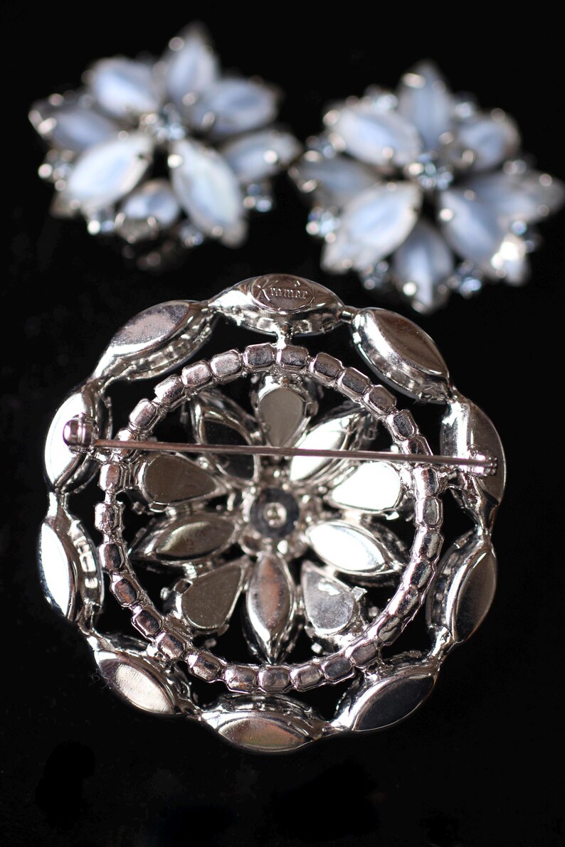 May include: A silver brooch with a floral design. The brooch is made of metal and has a clear, faceted stone in the center. The brooch is marked "mmma" on the back.