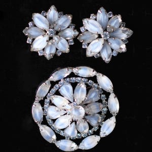 Könnte beinhalten: Ein Set aus drei Vintage-Schmuckstücken mit einer großen, runden Brosche und zwei passenden Ohrringen. Die Stücke sind aus silberfarbenem Metall gefertigt und haben einen Cluster aus weißen, irisierenden, ovalen Steinen.