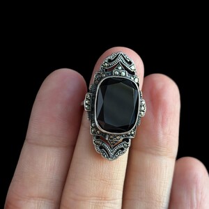 K&ouml;nnte beinhalten: Ein Vintage-Silberring mit einem gro&szlig;en, rechteckigen schwarzen Edelstein. Der Ring hat ein verziertes Design im Art-Deco-Stil mit kleineren, funkelnden Akzenten um den Hauptstein und auf den Schultern.