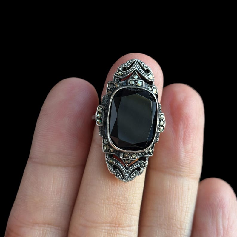 K&ouml;nnte beinhalten: Ein Vintage-Silberring mit einem gro&szlig;en, rechteckigen schwarzen Edelstein. Der Ring hat ein verziertes Design im Art-Deco-Stil mit kleineren, funkelnden Akzenten um den Hauptstein und auf den Schultern.