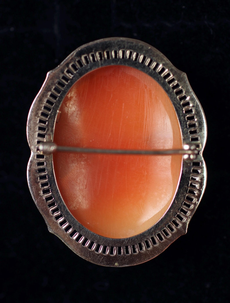 May include: A silver-toned brooch with a delicate, intricate design. The brooch features a large, oval-shaped coral cameo set in the center.