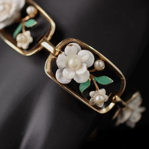 May include: A gold-tone bracelet with white flower accents and green leaves. The flowers are set against a black background.