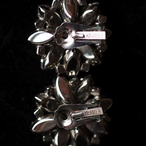 May include: A pair of silver tone earrings with a cluster of clear rhinestones. The earrings have a silver tone post and a silver tone butterfly clasp. The earrings are marked "Kramer" on the back.