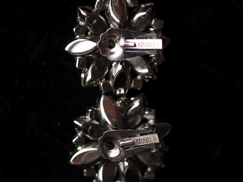 May include: A pair of silver tone earrings with a cluster of clear rhinestones. The earrings have a silver tone post and a silver tone butterfly clasp. The earrings are marked "Kramer" on the back.