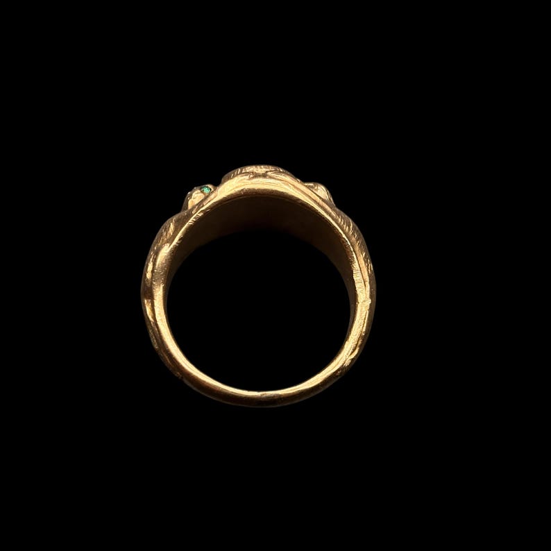 May include: A gold ring with a detailed design, featuring small green gemstones. The ring has an intricate pattern and is set against a black background, highlighting its ornate details.
