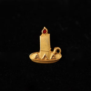 May include: A gold-colored pin with a candle design. The candle has a red gem for the flame and the letters "AAA" are inscribed on the base.