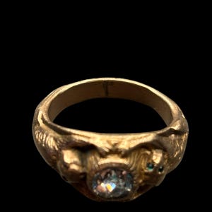 May include: A gold-colored ring with an oval, iridescent stone at the center. The ring features sculpted details of faces on either side of the stone and small green accents. The ring is set against a black background.
