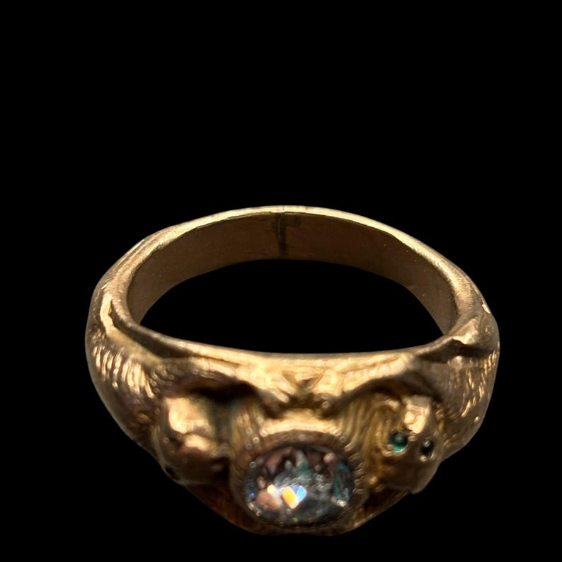 May include: A gold-colored ring with an oval, iridescent stone at the center. The ring features sculpted details of faces on either side of the stone and small green accents. The ring is set against a black background.