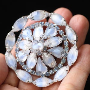 May include: A large, round, silver brooch with a cluster of white and blue rhinestones. The brooch is designed to look like a flower with a central white rhinestone.