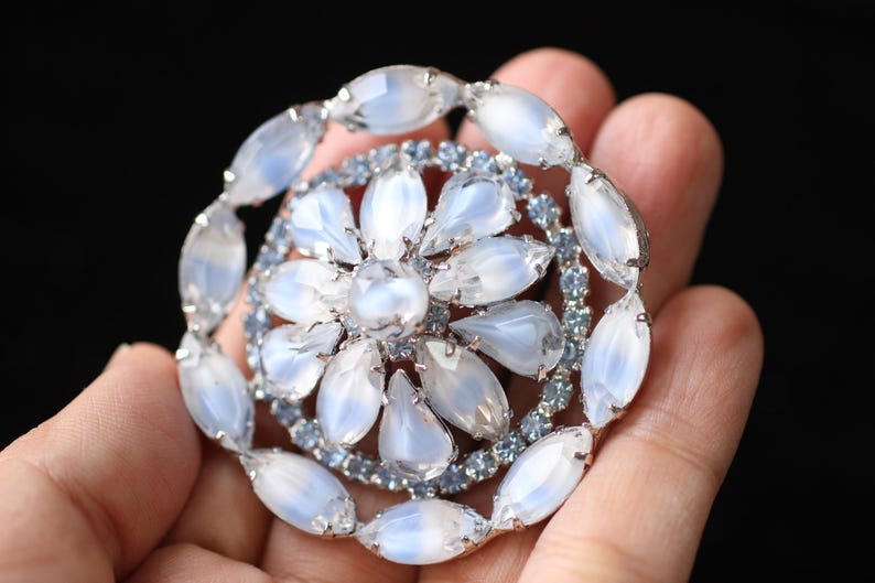 May include: A large, round, silver brooch with a cluster of white and blue rhinestones. The brooch is designed to look like a flower with a central white rhinestone.