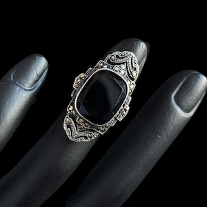 K&ouml;nnte beinhalten: Ein Vintage-Silberring mit einem gro&szlig;en, rechteckigen schwarzen Edelstein. Der Ring hat verzierte Details mit kleineren, funkelnden Steinen um den Hauptstein und an den Seiten. Der Ring wird auf einer schwarzen Oberfl&auml;che pr&auml;sentiert.