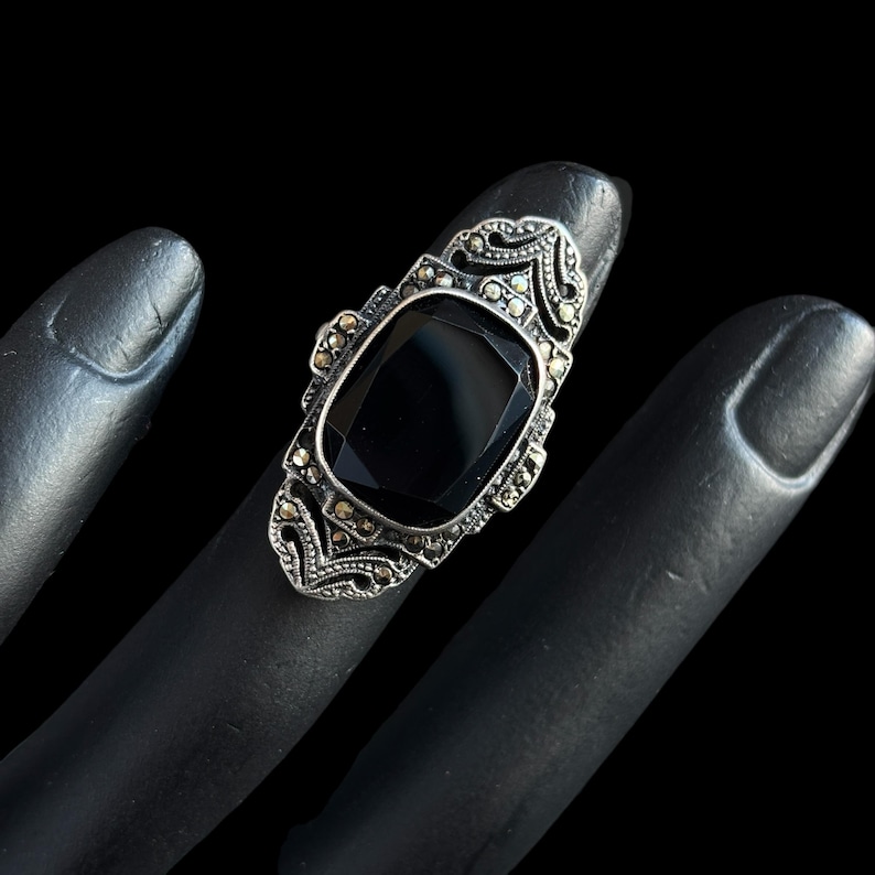 K&ouml;nnte beinhalten: Ein Vintage-Silberring mit einem gro&szlig;en, rechteckigen schwarzen Edelstein. Der Ring hat verzierte Details mit kleineren, funkelnden Steinen um den Hauptstein und an den Seiten. Der Ring wird auf einer schwarzen Oberfl&auml;che pr&auml;sentiert.